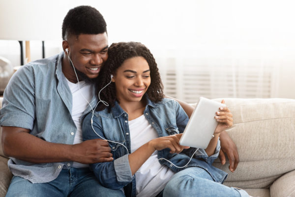 Positive African Couple Relaxing With Digital Tablet At Home, Sharing Earphones