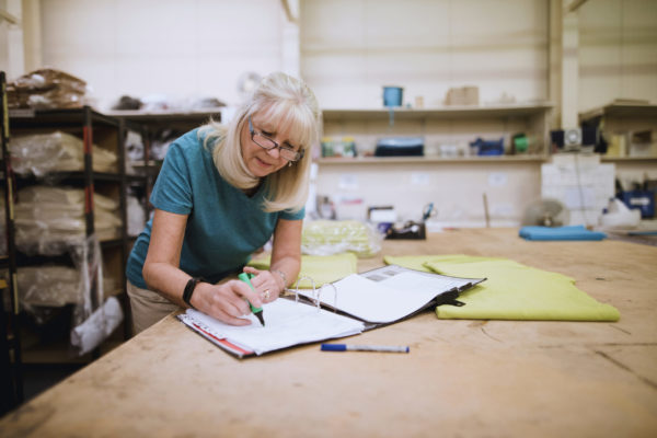 An older woman draws on a piece of paper on a binder in a factory