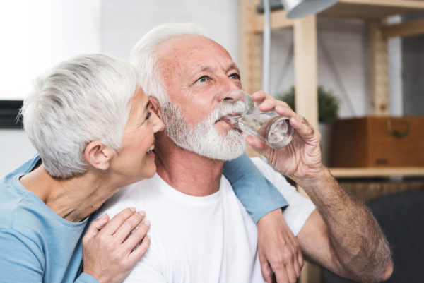 An older man drinks from a glass while his wife hugs him and smiles